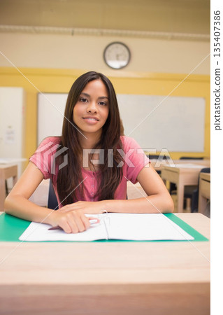 Female student sitting at wooden desk in classroom reviewing notes in open binder under clock 135407386