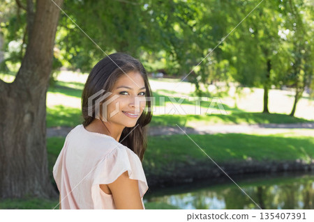 Asian woman standing beside calm pond in park bathing in dappled sunlight, copy space 135407391
