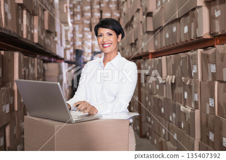 Asian woman working in warehouse aisle wearing button-down shirt at box workstation with laptop Asian woman working in warehouse aisle wearing button-down shirt at box workstation with laptop 135407392