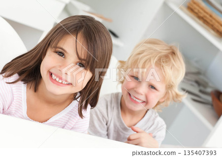 Brother and sister leaning over white countertop in kitchen with open shelving and wicker baskets 135407393