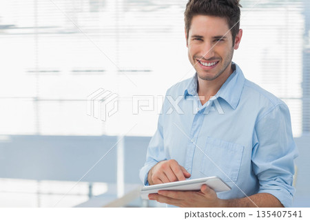 Man wearing light blue shirt rolling sleeves, smiling while holding tablet in office, copy space Man wearing light blue shirt rolling sleeves, smiling while holding tablet in office, copy space 135407541
