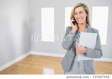 Middle-aged woman in suit taking smartphone call, holding pen, clipboard in empty room, copy space Middle-aged woman in suit taking smartphone call, holding pen, clipboard in empty room, copy space 135407548