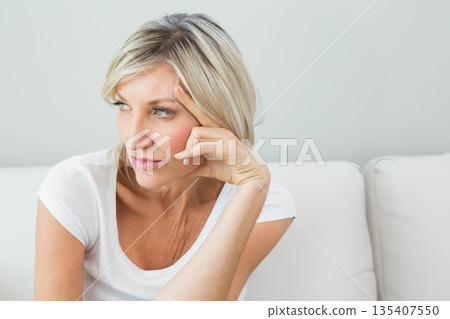 Sitting on white sofa in living room, woman wearing t-shirt with elbow on cushion, gazing sideways Sitting on white sofa in living room, woman wearing t-shirt with elbow on cushion, gazing sideways 135407550