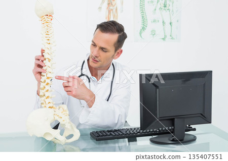 Mature doctor in lab coat pointing to vertebra model, sitting at desk with monitor and stethoscope Mature doctor in lab coat pointing to vertebra model, sitting at desk with monitor and stethoscope 135407551
