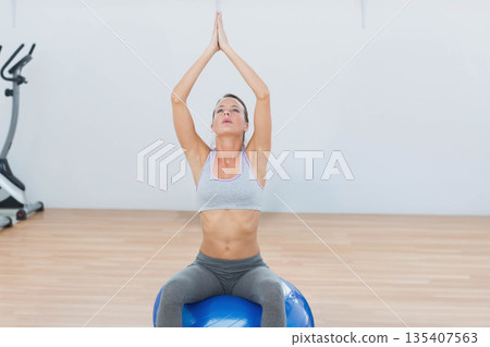 Woman in her 20s sitting on blue stability ball at gym pressing palms, stationary bike behind Woman in her 20s sitting on blue stability ball at gym pressing palms, stationary bike behind 135407563