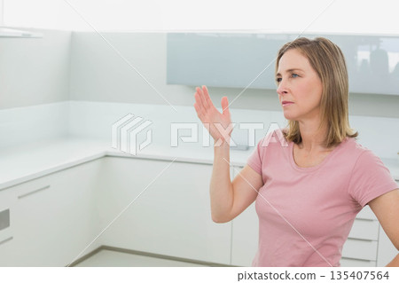 Mature adult female raising right hand in minimalist kitchen featuring white cabinets, copy space 135407564