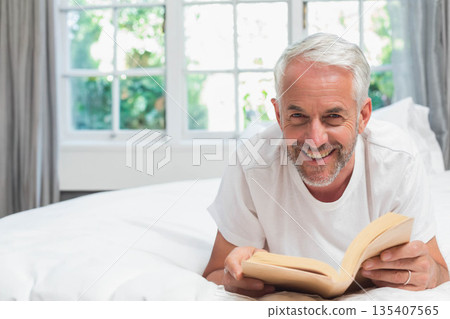 Senior man wearing white T-shirt lying on bed and reading paperback book in bedroom by window 135407565