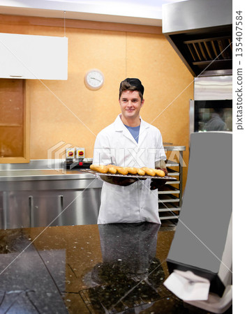 Male bakery worker wearing coat cap gloves holding bagel tray in bakery kitchen by prep table 135407584