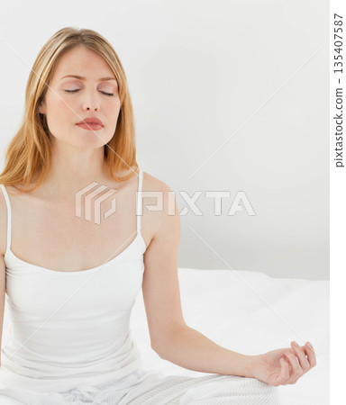 Woman in her 30s sitting cross-legged on bed in bedroom wearing striped pajama pants, copy space 135407587