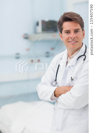 Male doctor with stethoscope standing arms crossed in exam room with bed and monitors, copy space 135407590