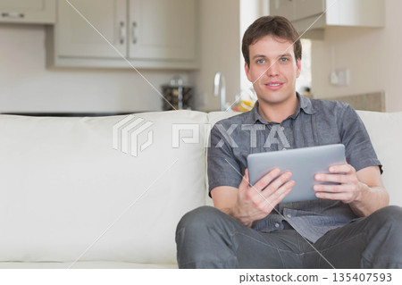 Mid adult man wearing grey shirt sitting on couch holding tablet with kitchen cabinets, copy space 135407593