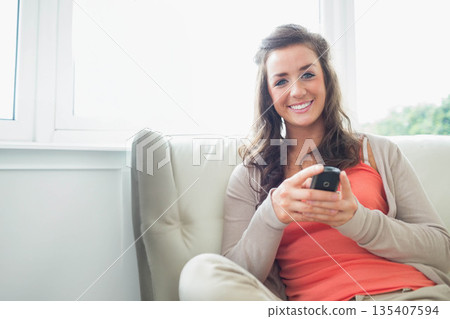 Woman holding smartphone while sitting on beige sofa by large window in living room, copy space 135407594