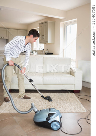 Man vacuuming area rug with vacuum cleaner on tiled floor in living room near kitchen cabinets 135407595