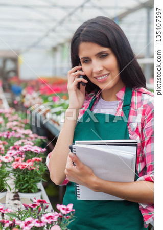 Female horticultural worker talking on smartphone while holding notebooks in greenhouse, copy space 135407597