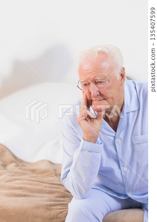 Senior man sitting on bed with blanket in pajamas resting on hand in bedroom, copy space 135407599