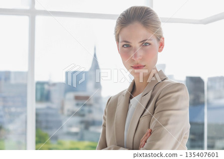 Businesswoman standing in office wearing blazer overlooking skyline by glass wall, copy space 135407601