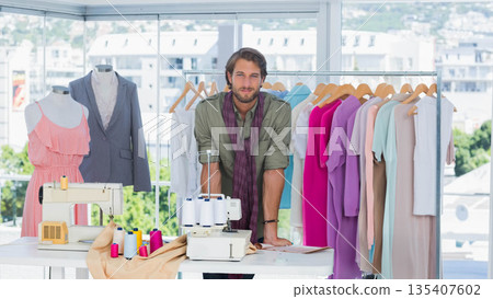 Male fashion designer leaning on white worktable in bright design studio with two sewing machines 135407602
