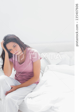 Woman sitting on bed with white sheets and pillows holding head and gazing downward 135407606
