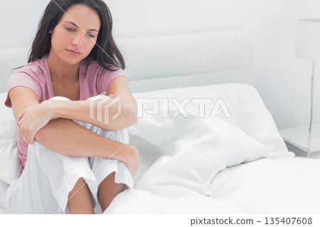 Woman sitting in pajamas on bed at home, looking downward at nightstand lamp, copy space Woman sitting in pajamas on bed at home, looking downward at nightstand lamp, copy space 135407608