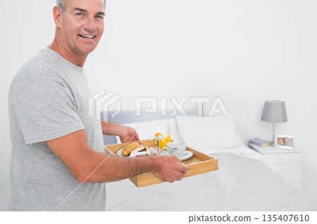Mature man holding wooden tray with croissant, orange juice beside made bed in bedroom, copy space 135407610