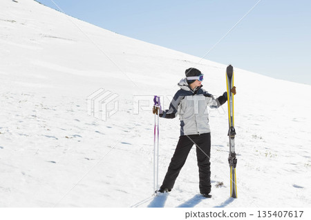 Skier standing on snowy hillside under clear blue sky, holding yellow skis and purple poles Skier standing on snowy hillside under clear blue sky, holding yellow skis and purple poles 135407617