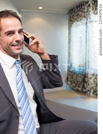 Man sitting on sofa at home in business suit holding smartphone with patterned curtains, copy space 135407619