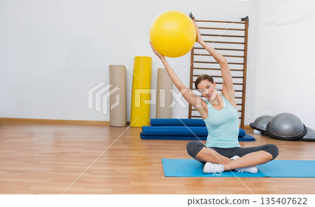 Woman lifting yellow exercise ball overhead sitting cross-legged on blue yoga mat in fitness studio 135407622