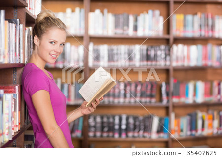 Woman wearing magenta top and jeans holding open paperback book in library aisle, copy space 135407625