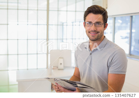 Office worker standing and smiling in office holding newspaper by glass partition, copy space Office worker standing and smiling in office holding newspaper by glass partition, copy space 135407627