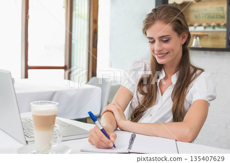Woman writing in spiral-bound notebook at coffee shop table with latte and laptop, copy space 135407629