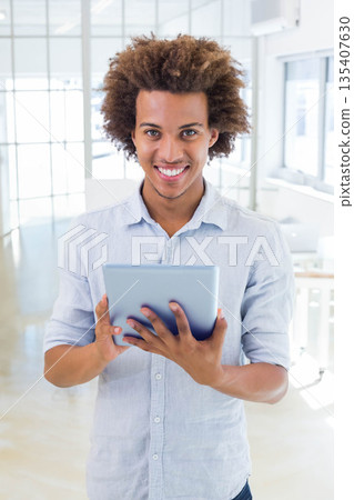 Man standing in modern office wearing light blue shirt holding tablet at chest level facing camera 135407630