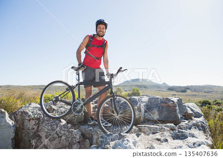 Male cyclist standing on rocky outcrop holding mountain bike wearing hydration backpack and helmet 135407636