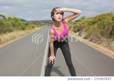 Woman wearing pink sports bra and black leggings leaning forward shielding eyes on rural paved road 135407637