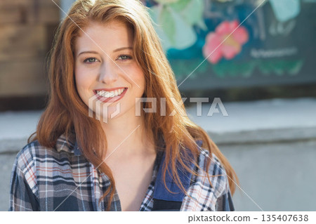Red-haired woman wearing blue white plaid shirt standing on patio with planter box and floral panel Red-haired woman wearing blue white plaid shirt standing on patio with planter box and floral panel 135407638