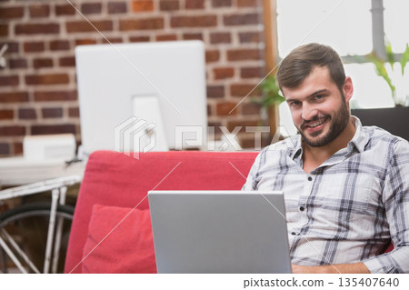 Man typing on laptop sitting on red sofa in loft workspace with monitor bicycle, copy space Man typing on laptop sitting on red sofa in loft workspace with monitor bicycle, copy space 135407640
