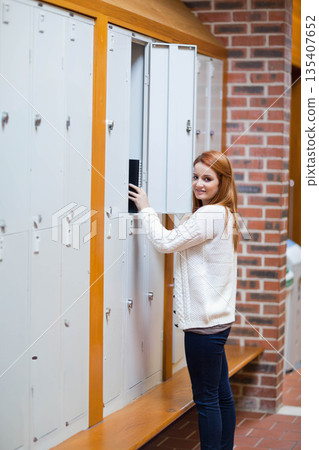 Teenage female student opening locker and retrieving black binder in school corridor, copy space 135407652