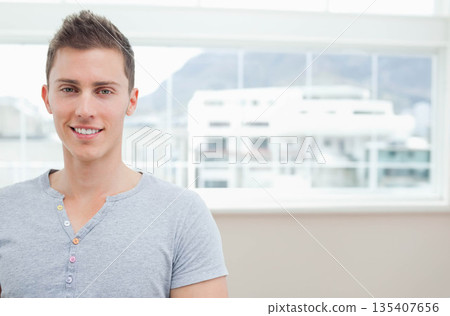 Man standing by window frame in apartment wearing button-neck tee overlooking cityscape, copy space Man standing by window frame in apartment wearing button-neck tee overlooking cityscape, copy space 135407656