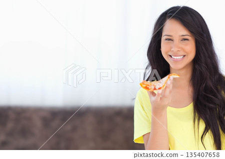 Woman holding cheese pizza slice near mouth on sofa at home wearing yellow T-shirt, copy space 135407658