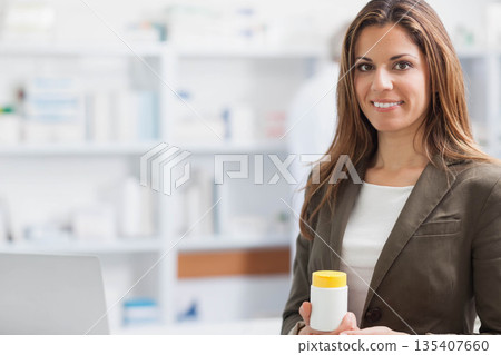 Female pharmacist smiling, holding white medication container at counter with laptop, copy space 135407660