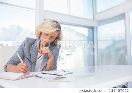 Woman in blazer writing in notebook holding glasses at office desk with mountain view, copy space Woman in blazer writing in notebook holding glasses at office desk with mountain view, copy space 135407662
