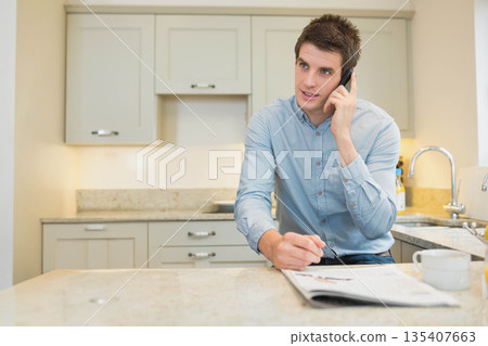 Man using smartphone while reviewing printed pages on kitchen island with coffee mug, copy space Man using smartphone while reviewing printed pages on kitchen island with coffee mug, copy space 135407663