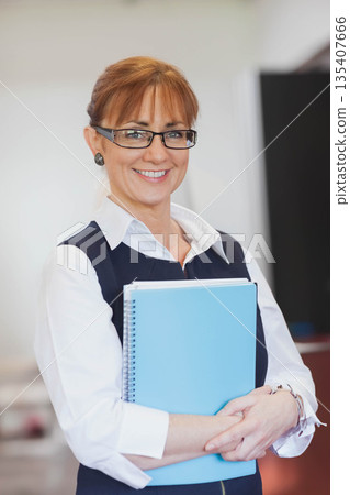 Professional woman holding blue binder and smiling in office wearing business attire Professional woman holding blue binder and smiling in office wearing business attire 135407666