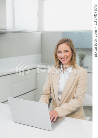 Senior woman wearing beige blazer smiling, typing on silver laptop at white office desk, copy space Senior woman wearing beige blazer smiling, typing on silver laptop at white office desk, copy space 135407678