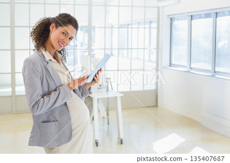 Pregnant businesswoman standing at office wearing grey blazer holding tablet near desk, copy space 135407687