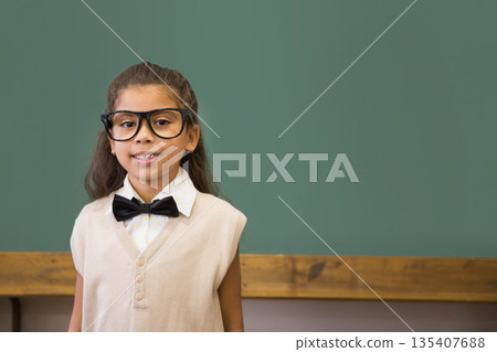 Hispanic child standing at chalkboard chalk tray in classroom wearing eyeglasses bowtie, copy space Hispanic child standing at chalkboard chalk tray in classroom wearing eyeglasses bowtie, copy space 135407688