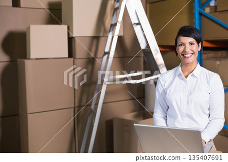 Woman in white shirt standing at warehouse desk using laptop amid boxes and shelving, copy space 135407691