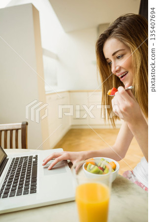 Woman in twenties eating fruit from bowl while working on laptop at kitchen table, copy space Woman in twenties eating fruit from bowl while working on laptop at kitchen table, copy space 135407694