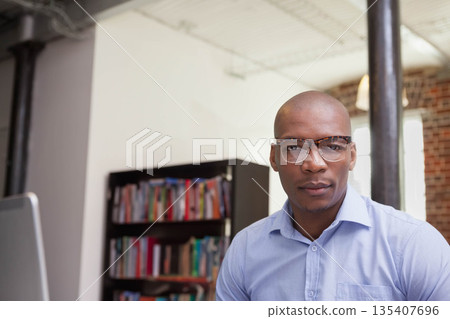 African American man in shirt adjusting eyeglasses leaning on office desk by bookshelf, copy space 135407696