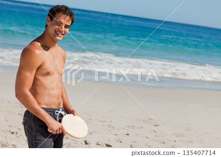 Shirtless man wearing swim trunks holding wooden paddle and standing on shore by waves, copy space Shirtless man wearing swim trunks holding wooden paddle and standing on shore by waves, copy space 135407714