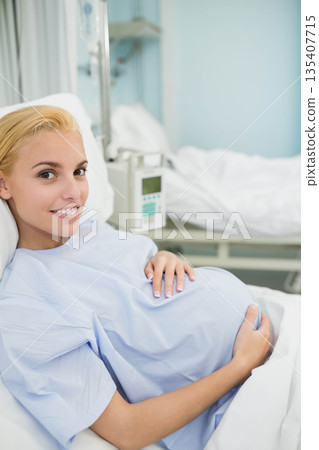 Pregnant woman holding belly reclining on hospital bed wearing gown beside IV stand 135407715